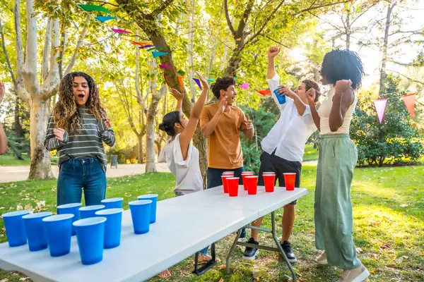 Bir grup mutlu çoklu etnik arkadaş parkta bir partide bira pong oynayarak kutlama yapıyor.