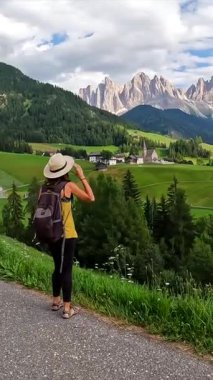 Bayan turist şapkasını tutarken arka planda küçük dağlarla birlikte Val di Funes 'deki St. Magdalena kilisesine bakıyor.