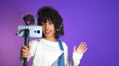 Dijital içerik yaratan, mikrofona tutkulu bir şekilde el sallayan ve konuşan kendine güvenen kadın etkili, canlı mor arka plan üzerine video blogu kaydediyor.