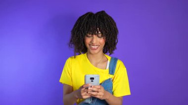 Genç Afro-Amerikalı kadın akıllı telefonuyla uğraşırken gülümsüyor, kot pantolon giyiyor ve parlak mor arka planda sarı gömlek giyiyor.