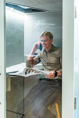 Gülümseyen yönetici, iş yerindeki şeffaf kabinde belgeleri okuyor ve cep telefonuyla konuşuyor.