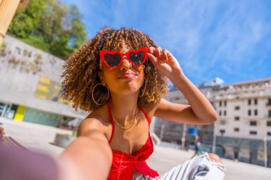 Personal perspective of a cool latin young and beauty woman with curly hair wearing hear shape sunglasses taking selfie and joking in the city