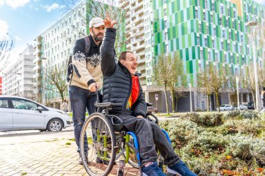 Bakıcı tekerlekli sandalyedeki mutlu engelli adamı itiyor, şehir ortamında el sallıyor ve gülümsüyor.