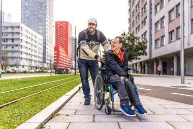 Bakıcı, tekerlekli sandalyeli gülümseyen engelli bir adamı hareketli bir şehir kaldırımında iterken bir anlık dostluğun ve desteğin tadını çıkarıyor.