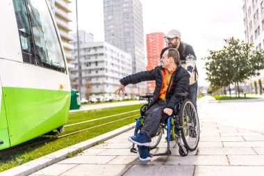Tekerlekli sandalyedeki mutlu engelli adama yardım eden bakıcı şehir merkezinde modern bir tramvaya uzanıyor.
