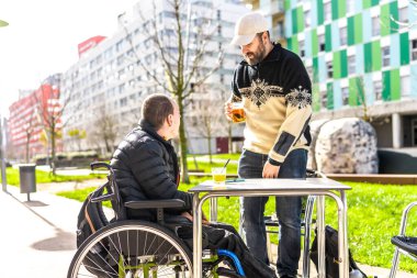 İki arkadaş açık hava masasında içmekten zevk alıyor. Biri tekerlekli sandalye kullanıyor. İlişki ve dostluğu gelişigüzel gösteriyor.