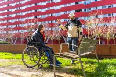 Tekerlekli sandalyedeki genç adam, şehir parkında bir arkadaşını selamlıyor, dostluk, destek ve kapsama gösteriyor.