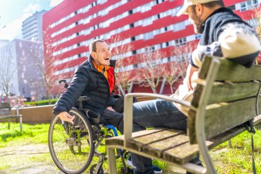 Tekerlekli sandalyedeki genç adam şehir parkında müzisyen arkadaşıyla gülüp eğleniyor.