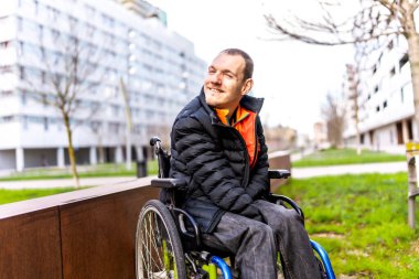 Tekerlekli sandalye kullanan genç adam, şehir parkında eğlenirken gülümsüyor, dahil olma ve erişilebilirliği teşvik ediyor.