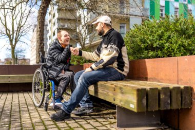 Biri tekerlekli sandalyede otururken iki arkadaş el sıkışıyor, ikisi de canlı şehir parkında güneşli bir günün tadını çıkarıyor.