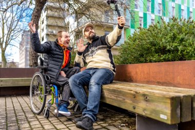 İki neşeli içerikli yaratıcı, biri tekerlekli sandalyede, bir şehir parkında vibratör kullanarak bir video blogu kaydediyor.
