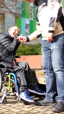 Tekerlekli sandalyedeki erkek kullanıcı ve arkadaşı sıcak bir kahkaha paylaşıyor, güneşli bir günde kentteki bankta otururken gerçek dostluk ve duygusal bağ yayıyor.
