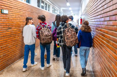 Sınıflar arası koridorda yürüyen, sırt çantaları taşıyan ve sohbet eden çeşitli öğrenci gruplarının arka görüntüsü.