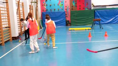 Turuncu ve mor tişörtler giyen ilkokul öğrencileri beden dersinde florbol oynuyorlar.