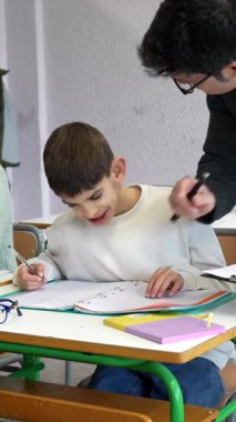 Yardımcı öğretmen, bir sınıf aktivitesi sırasında Down sendromlu genç bir öğrenciye yardımcı oluyor.