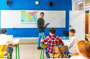 Öğrenciler dizüstü bilgisayarlar ve kitaplarla çalışırken coğrafya öğretmeni sınıfta yürüyor