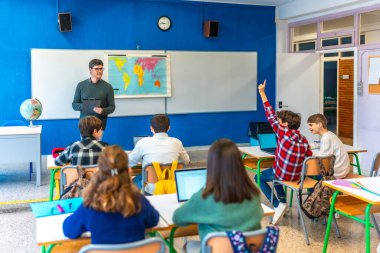 Bir grup genç öğrenci el kaldırıyor ve sınıfta öğretmen ve dizüstü bilgisayarlarla coğrafya dersini takip ediyor.