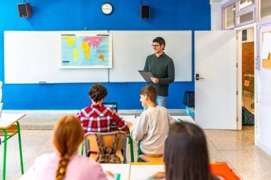 Coğrafya öğretmeni elinde bir not panosu olan bir dersi anlatıyor ve sınıfta ilkokul öğrencilerine gülümsüyor.