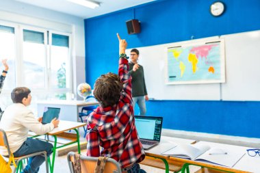 İlkokul öğrencisi coğrafya dersinde bir soruyu cevaplamak için elini kaldırırken, arka planda öğretmen ve sınıf arkadaşları vardı.