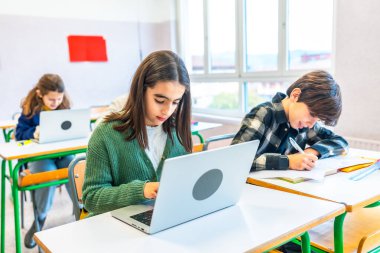 İlkokul öğrencileri sınıf aktivitelerine katılıyorlar, araştırma için dizüstü bilgisayar kullanıyorlar ve dersler sırasında özenle not alıyorlar.