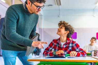 İlkokul öğretmeni gülümseyen bir öğrenciyle meşgul olurken aynı zamanda masalarında neşeli bir sınıf ortamında ders veriyor.