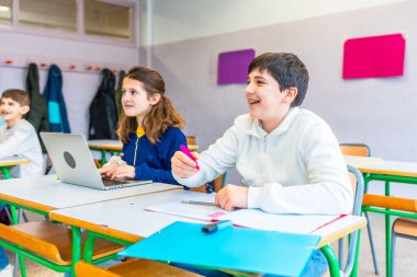 İlkokul öğrencileri eğitim faaliyetlerinde bulunuyorlar, teknolojiden faydalanıyorlar ve parlak renkli bir sınıf ortamında sınıf arkadaşlarıyla işbirliği yapıyorlar.