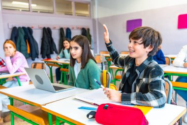 Sınıf arkadaşları laptoplarını kullanırken ve öğretmenlerini dinlerken ders sırasında soruları cevaplamak için sınıfta elini kaldıran zeki çocuk.