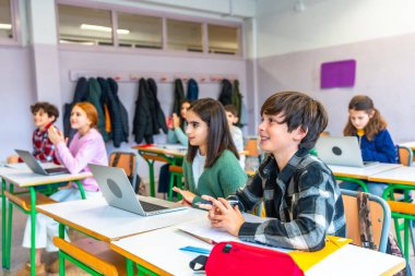 İlkokul öğrencileri öğretmeni dinliyor ve modern bir sınıftaki dizüstü bilgisayarlarla derse katılıyorlar.