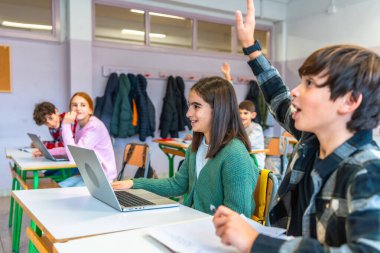 İlkokul öğrencileri etkileşimli bir sınıfta el kaldırıyor ve dizüstü bilgisayar kullanıyor, modern eğitim faaliyetlerine katılıyorlar.