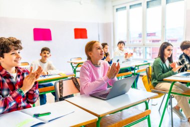 Bir grup ilkokul öğrencisi modern bir sınıfta el çırpıyor, bazıları dizüstü bilgisayar kullanıyor, bir ders sırasında bağlılık ve takdir gösteriyor