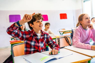 İlkokul öğrencisi ders sırasında bir soruyu cevaplamak için el kaldırıyor, öğrenme ve sınıfa katılma hevesini gösteriyor.