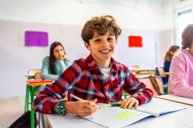 Mutlu ilkokul öğrencisi sınıfta gülümseyip not defterine yazarken sınıf arkadaşları arka planda.