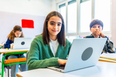 İlkokul öğrencisi sınıfta dizüstü bilgisayar kullanıyor, gülümsüyor ve arka planda sınıf arkadaşları ile kameraya bakıyor.