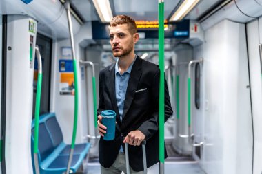 İşe metroyla gidip gelen genç bir işadamı, elinde yeniden kullanılabilir bir kahve fincanı ve pencereden dışarı bakarken bir poşet tutuyor.