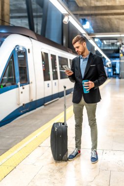 İş adamı metro istasyonunda duruyor, akıllı telefon kullanıyor, elinde kahve tutuyor, tramvayı ile treni bekliyor.