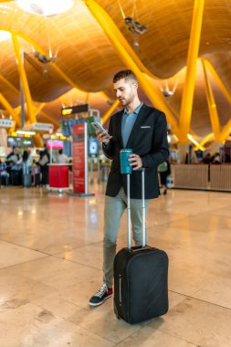 Havaalanında bekleyen bir iş adamı akıllı telefonunu kullanıyor ve elinde yeniden kullanılabilir bir kahve fincanı tutuyor.