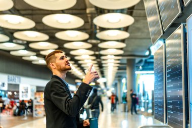 İş adamı akıllı telefonuna danışıyor ve havaalanındaki ekranlarda gösterilen uçuş bilgilerini kontrol ediyor.