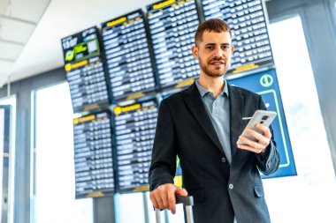 İş adamı bavulu tutuyor ve havaalanından kalkmayı beklerken akıllı telefon kullanıyor.
