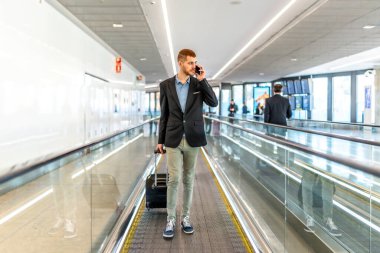 İş adamı bavulu çekiyor ve havaalanında yürürken akıllı telefondan konuşuyor.