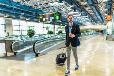Havaalanı terminalinde yürüyen bir iş adamı, akıllı telefon, seyahat ve teknolojiyi harmanlarken bir bavulu çekiyor.