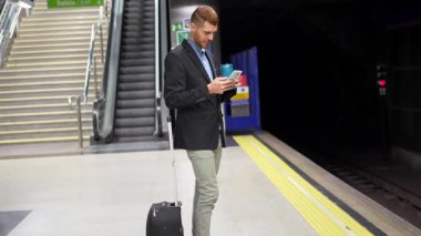 Kafkasyalı genç işadamı metroyu bekliyor, tekrar kullanılabilir şişeden içiyor ve akıllı telefonunu kontrol ediyor.