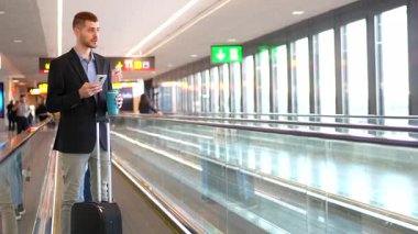 Genç işadamı, havaalanında yürürken akıllı telefon kullanıyor ve kahve içiyor.