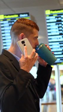 Genç işadamı kahve içiyor ve havaalanında cep telefonu kullanıyor, kalkış panosundaki uçuş bilgilerini kontrol ediyor.