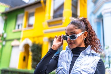 Modern fütüristik kıyafetler ve güneş gözlüğü takan Afrikalı Amerikalı kadın renkli bir sokakta duruyor.