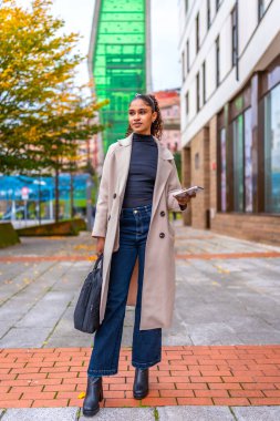 Şık bir Afrikalı iş kadınının dikey portresi. Şehirde elinde akıllı telefon ve çanta duruyor.