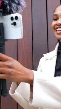 Afro-Amerikalı genç kadın içerik yaratıcısı şehirdeki telefon kullanarak yeni bir video bloğu kaydediyor.
