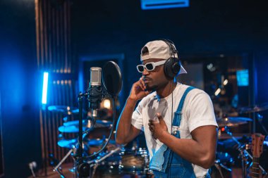 Genç, şık erkek sanatçı, modern ekipman ve canlı ışıklarla çevrili profesyonel bir müzik stüdyosunda tutkuyla şarkı kaydediyor.