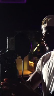 Müzik yapımında yetenekli bir sanatçı, kulaklıkları ayarlıyor ve mikrofona şarkı söylüyor, mor stüdyo ışıklarıyla aydınlatılıyor.