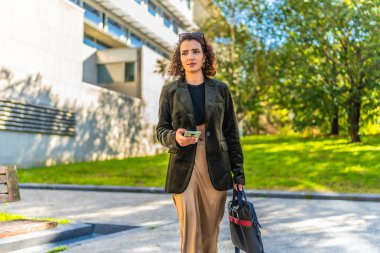 Genç Arap girişimci dizüstü bilgisayar çantası ve cep telefonu taşıyan ağaçlarla cadde boyunca yürüyor