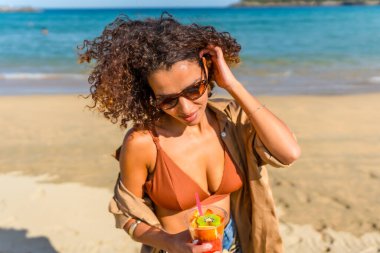 Yaz kıyafetleri içinde tatlı bir Arap kadın sahilde elinde taze bir bardak tropikal meyve tutuyor.
